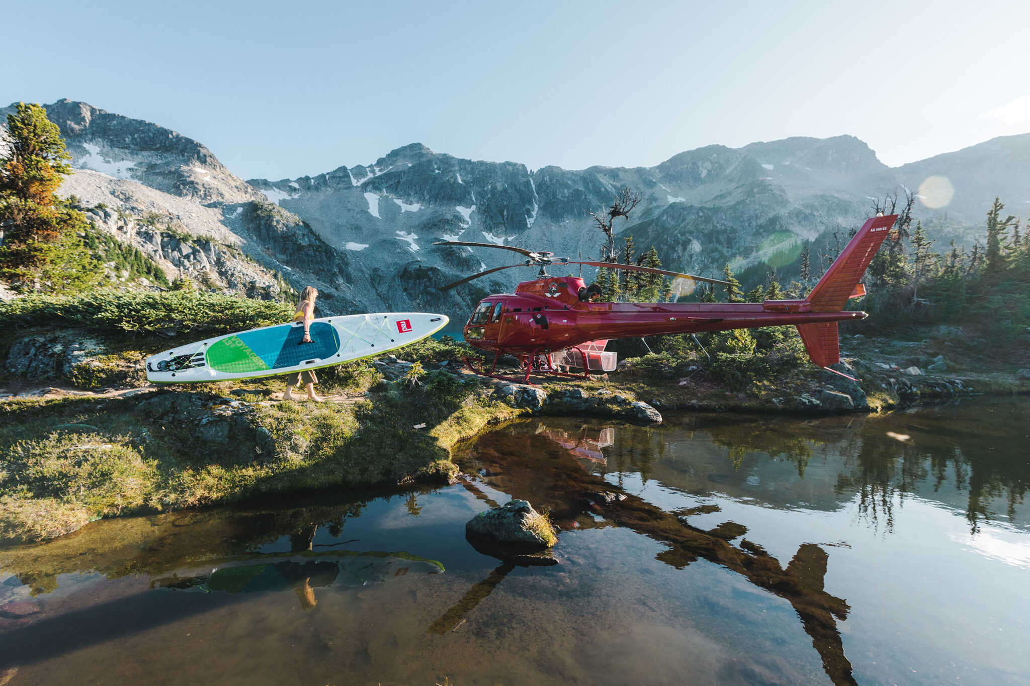 A helicopter is sat by a beautiful mountain lake as a woman gets a paddleboard out.