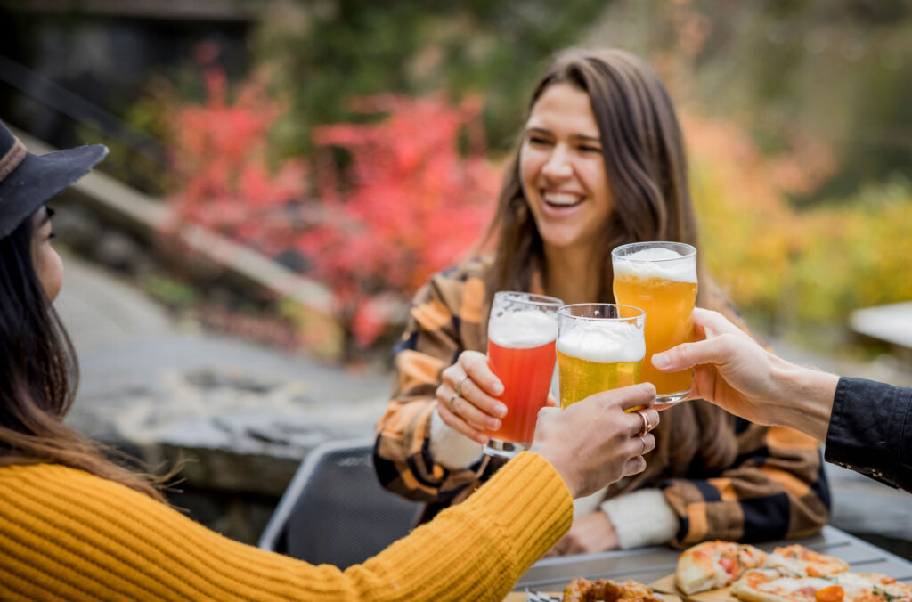Friends say cheers with their beers on a patio in Whistler with the beautiful fall colours in the background.