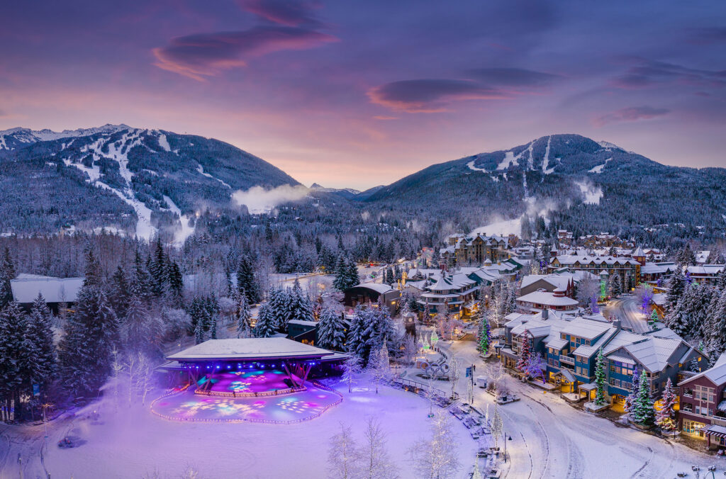 A drone shot of Whistler Village as the sun sets creating a purple glow over the lights of Whistler Village with Whistler and Blackcomb mountains in the background.