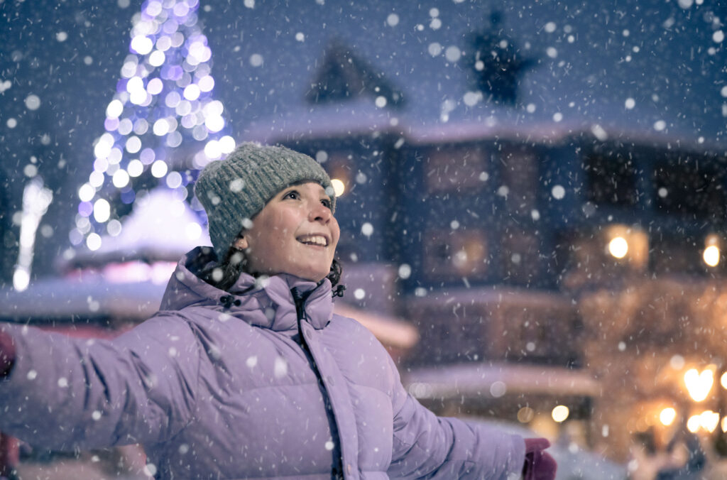 A young girl spins in the snow as it falls in Whistler Village with sparkling, festive lights behind her.