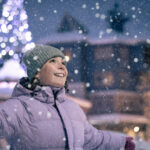 A young girl spins in the snow as it falls in Whistler Village with sparkling, festive lights behind her.