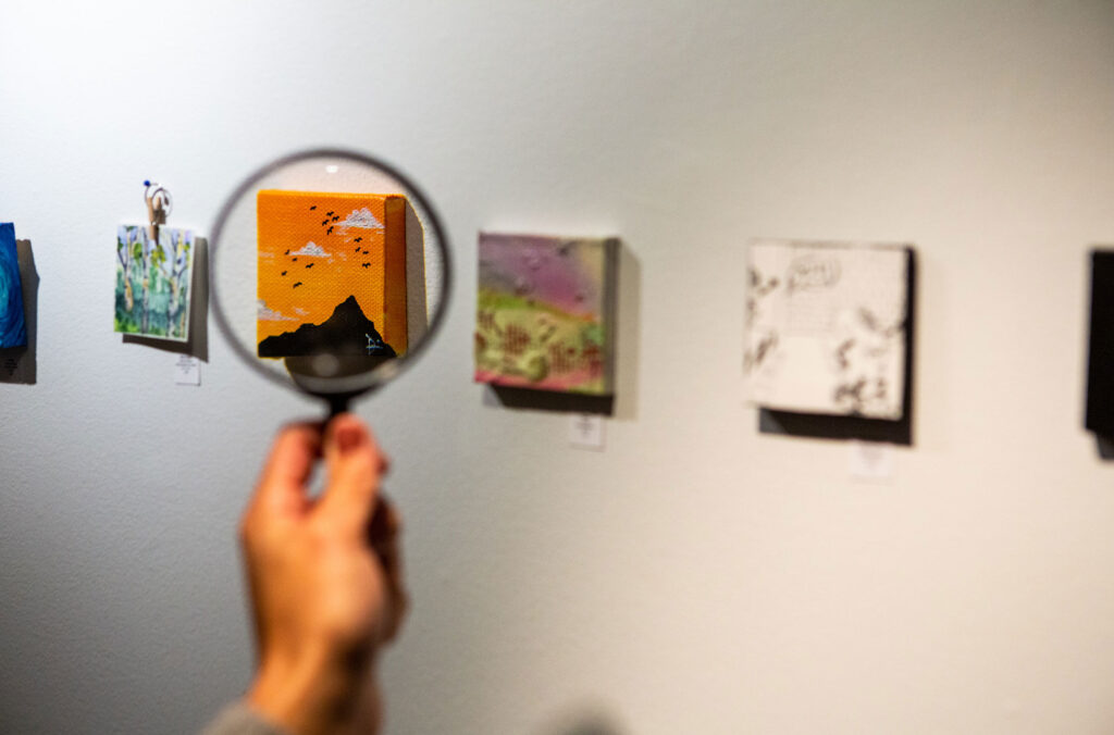 A person holds a magnifying glass up to the artwork at the Teeny Tiny Art Show in Whistler.