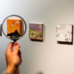 A person holds a magnifying glass up to the artwork at the Teeny Tiny Art Show in Whistler.