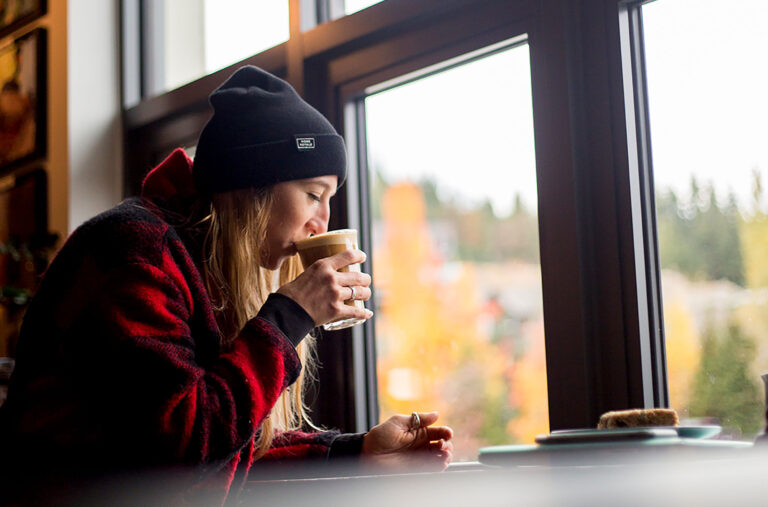 A woman drinks a coffee overlooking Blackcomb Mountain at Cranked Coffee in Rainbow, Whistler.