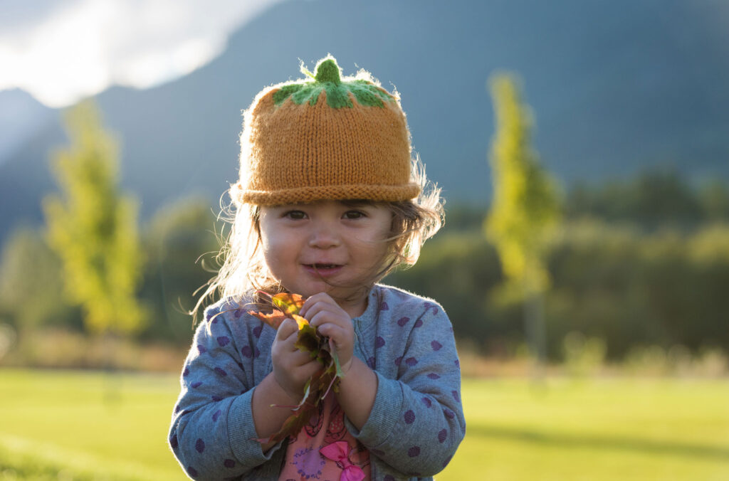 A young child looks at the camera holding fall leaves in her hands. She's wearing a cute, pumpkin hat.