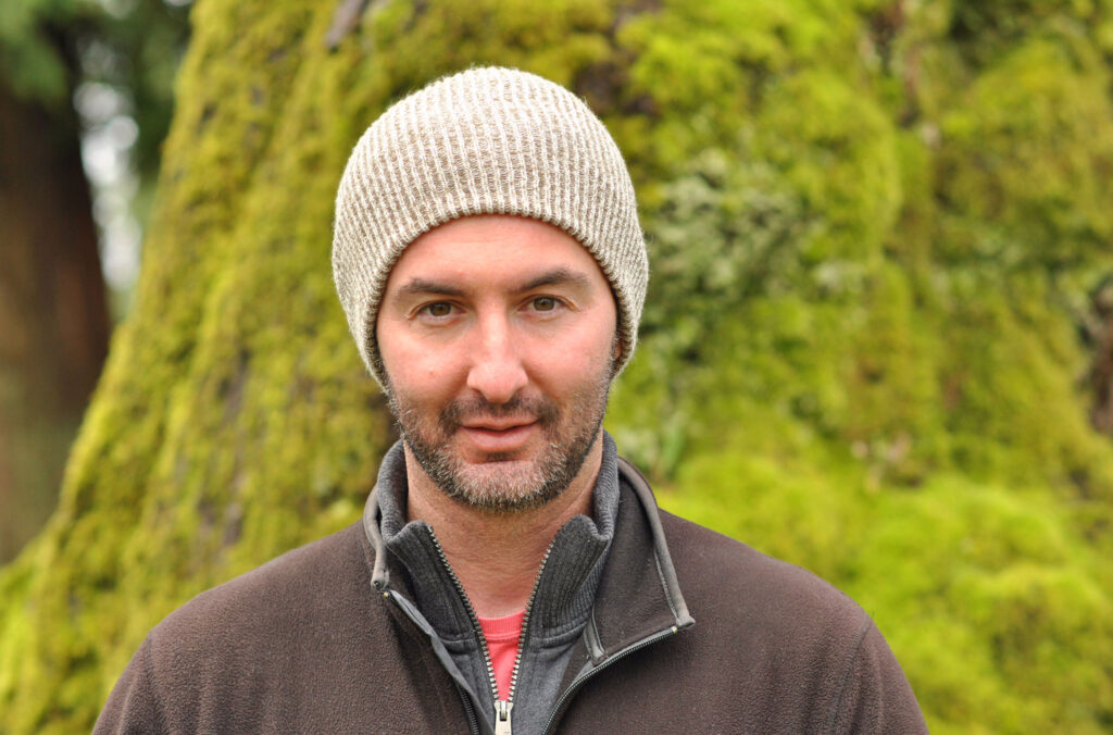 A picture of Ira Pettle looking directly at the camera stood in a lush forest.