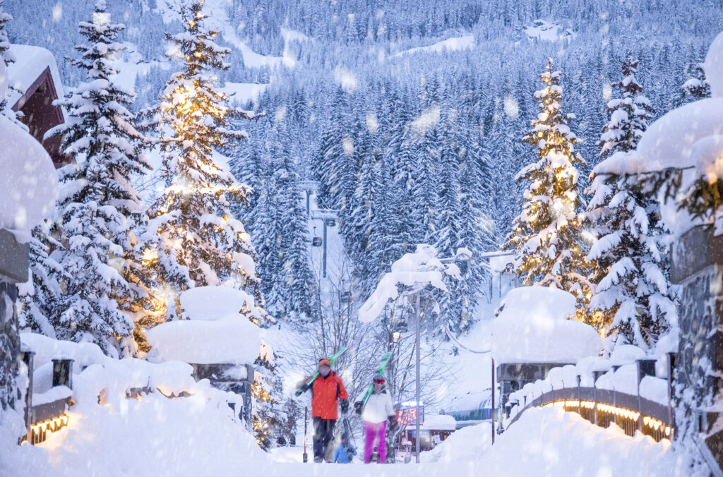 Two skiers walk over the bridge at the base of Creekside. The gondola goes up behind them.