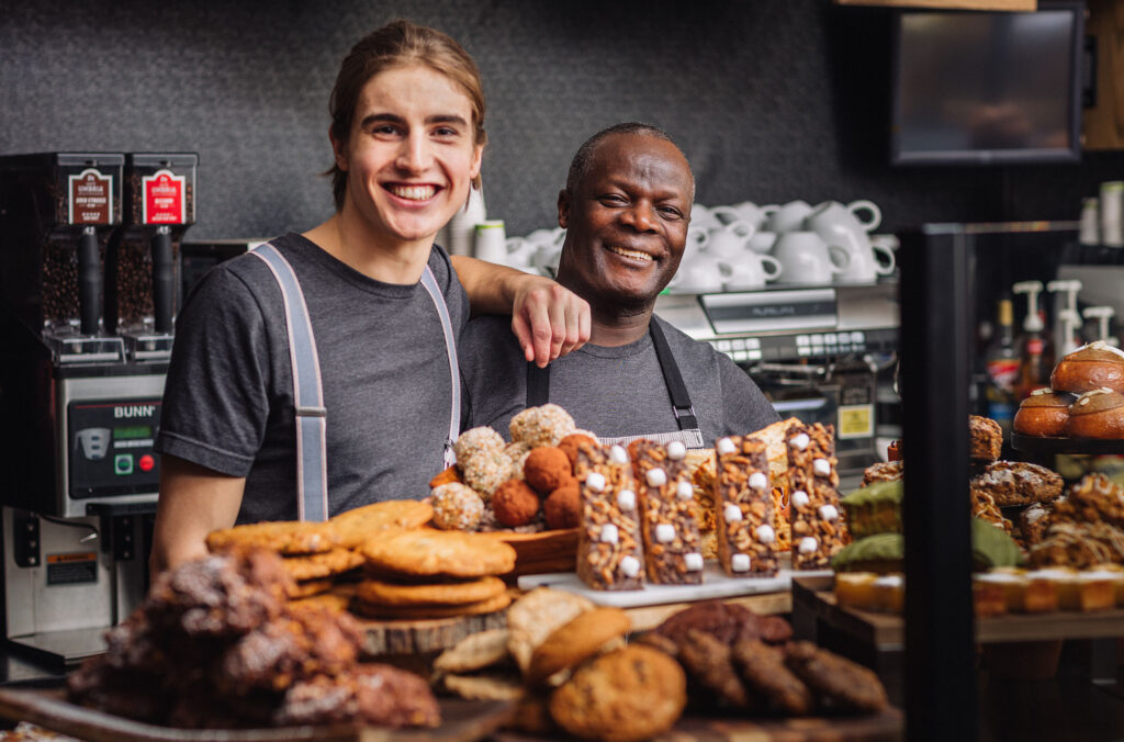 Two work colleagues at Portobello in Whistler stand behind a counter packed with delicious-looking sweet treats.