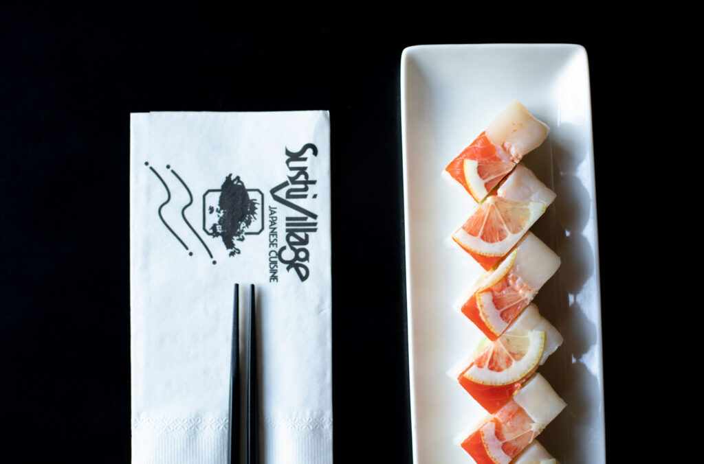 A plate with one of Sushi Village's famous sushi rolls on it next to a napkin with chopsticks.