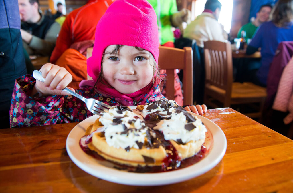A child smiles as she tucks into a huge waffle topped with whipped cream and chocolate sprinkles at the Crystal Hut at Whistler Blackcomb.