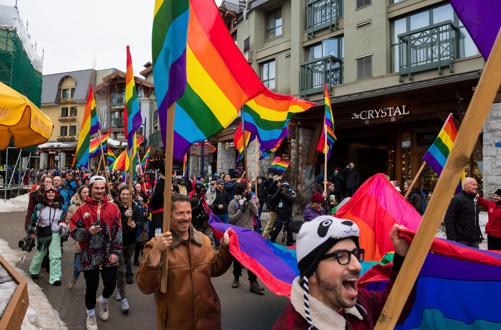 The Whistler Pride and Ski Festival parade makes its way through Whistler Village to Olympic Plaza. People are carrying rainbow flags and banners.