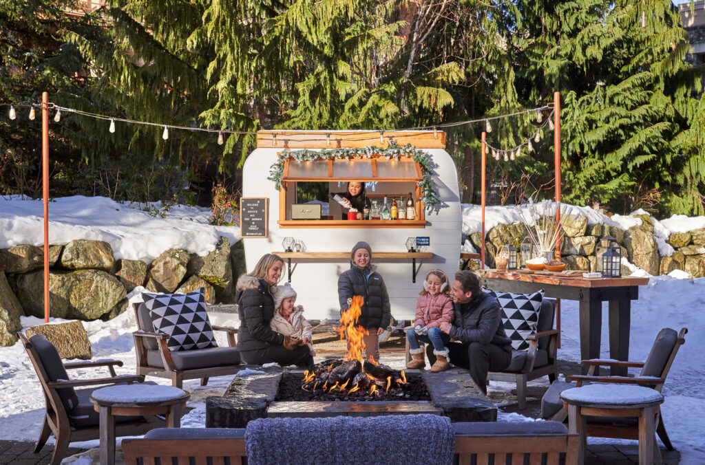 A family with three, young kids sit around the fire on the Braidwood Tavern's patio roasting marshmallows in front of the vintage camper with a server adding whipped cream to hot chocolates.