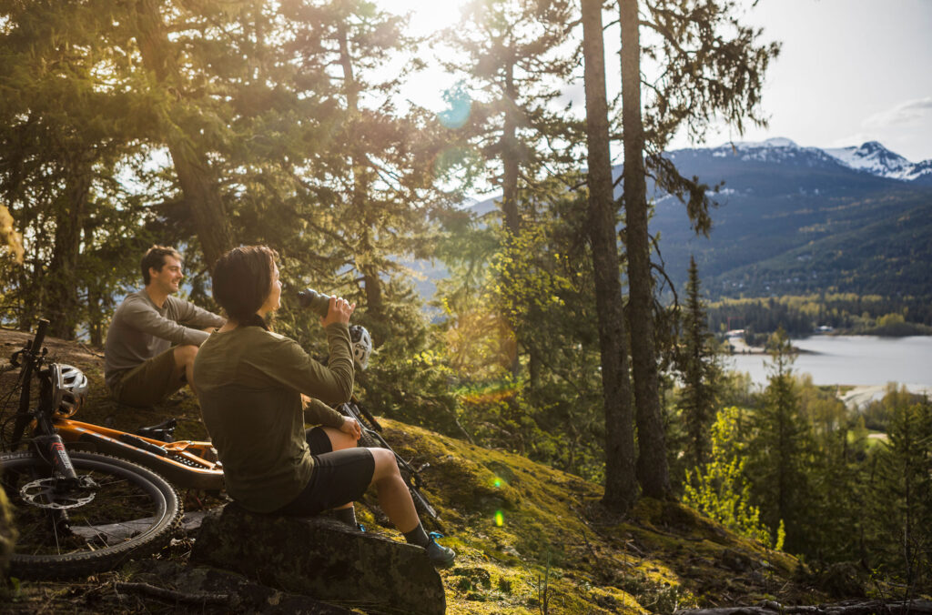 Two bikers sit on a hilly outcrop after a bike ride in the Whistler forest and enjoy views out over a lake.