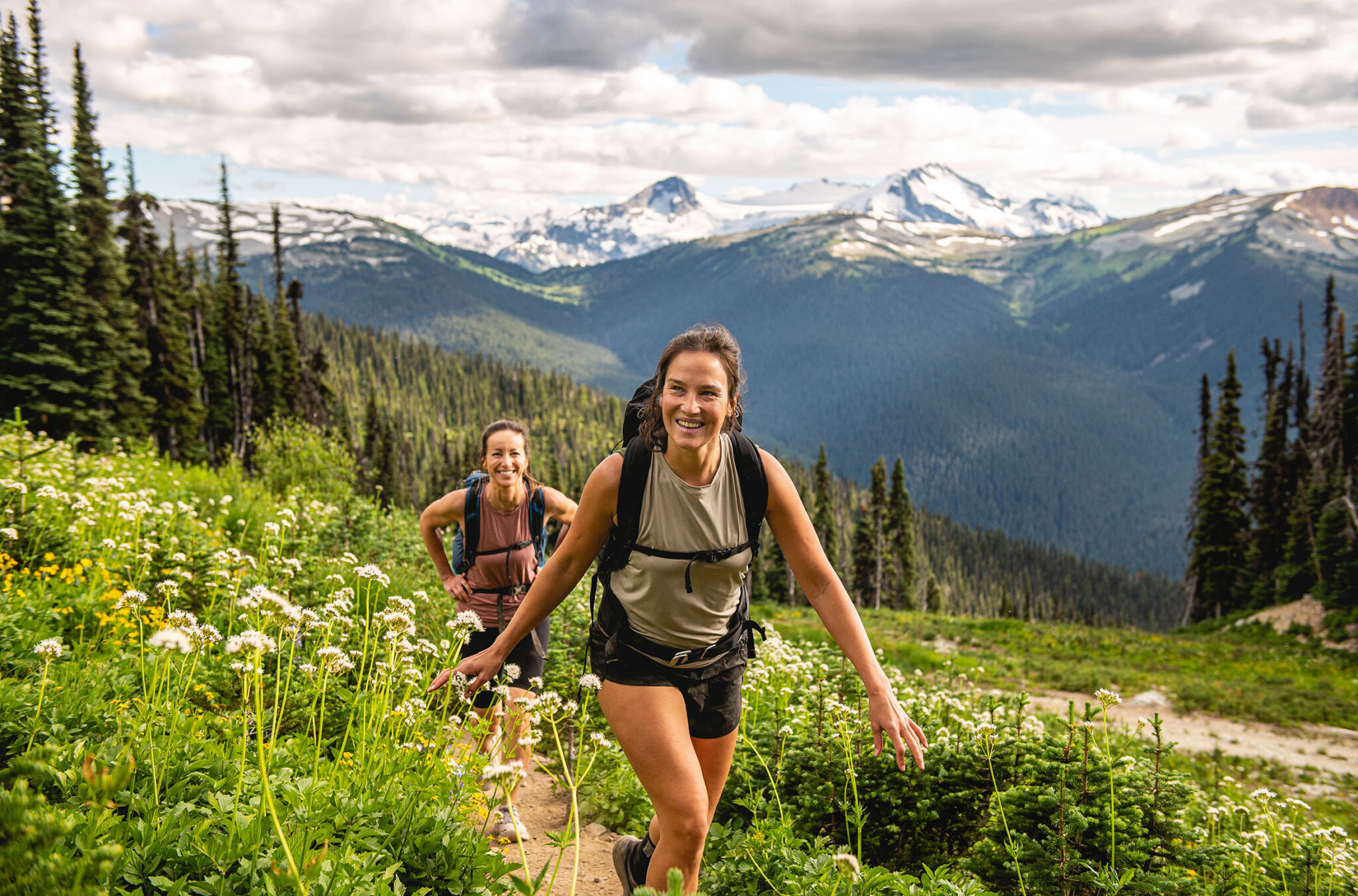 Two women hike in Whistler's alpine in the summer, there are wildflowers blooming around them.