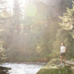 A woman stands in the sun on the bank of a river with the lush forest behind her in Whistler.