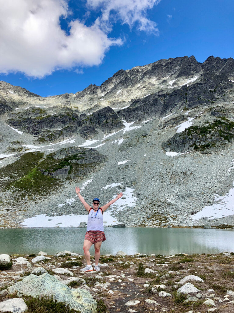 Kate Heskett waving to the camera stood in front of an alpine lake on Whistler Blackcomb.