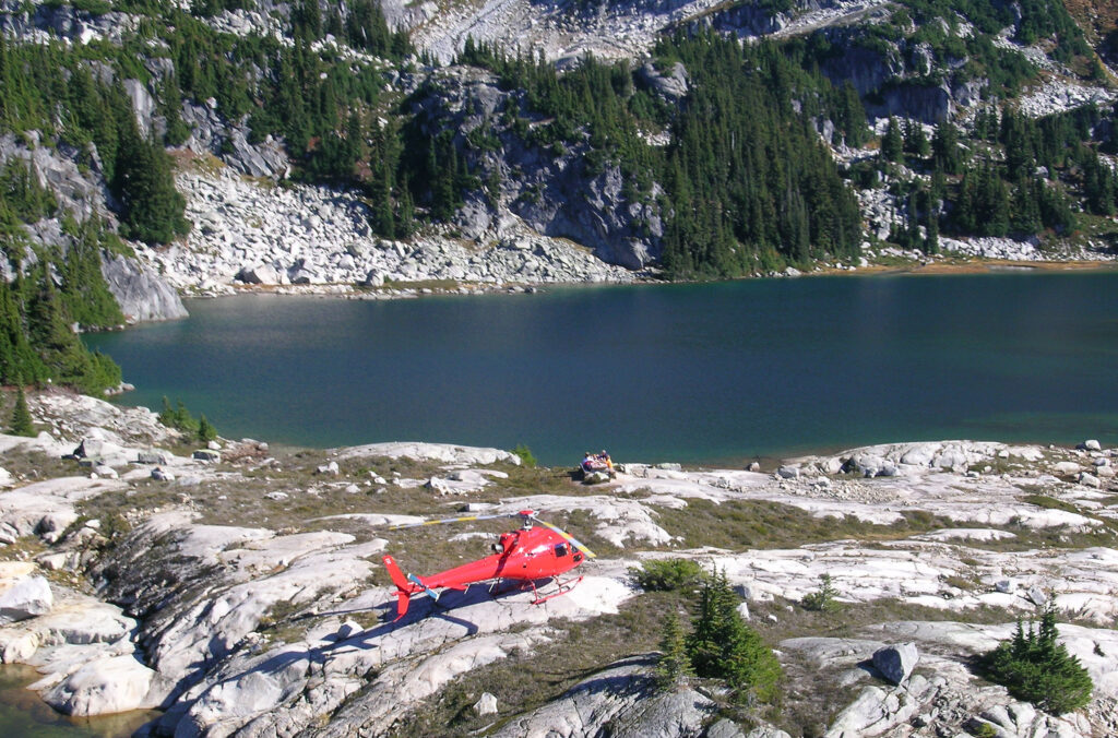 A red helicopter by a secluded alpine lake with a small picnic table on the edge of the water.