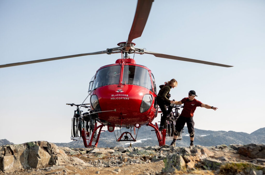 One of the bikers gets out of a red, Blackcomb Helicopter on top of a mountain, with the pilot pointing to where they need to go.