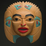 An Indigenous mask carving showing a woman's face with salmon in the design, coloured natural wood, teal, red and black.