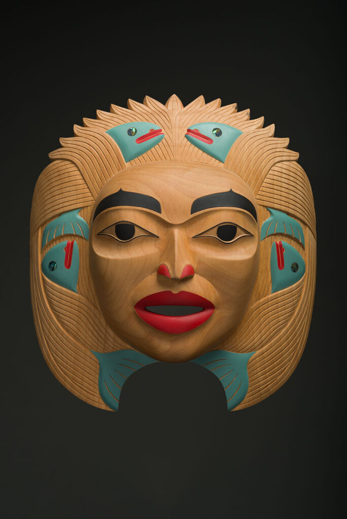 An Indigenous mask carving showing a woman's face with salmon in the design, coloured natural wood, teal, red and black.