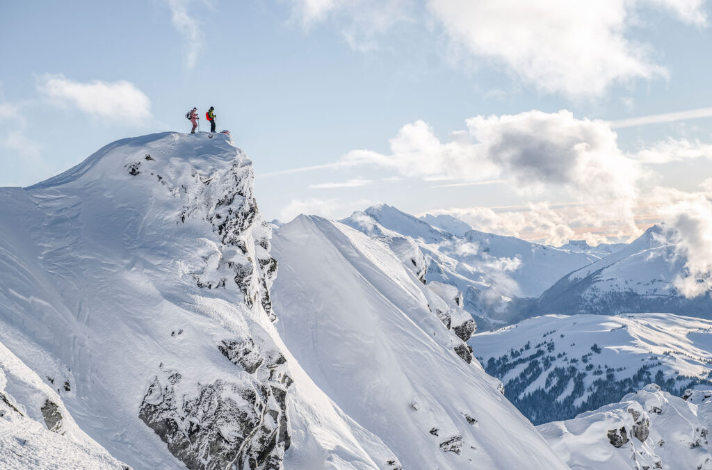 Two explorers stand on top of a snowy mountain looking out over the Coast Mountains.