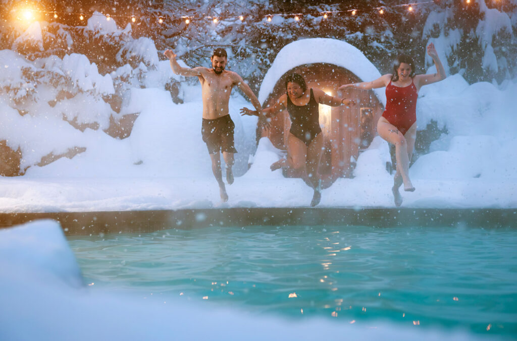 Three friends jump into a heated pool which is surrounded by snow, lit by fairy lights and the glow from a barrel sauna. 