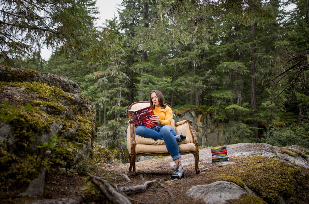 A woman sits on a padded chair set on a rock outside in the forests of Whistler. She reads from a stack of books.