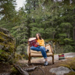 A woman sits on a padded chair set on a rock outside in the forests of Whistler. She reads from a stack of books.