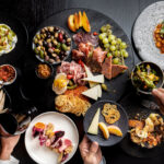 A beautiful and colourful array of food laid out on a dark table with multiple hands reaching in to take a bite.