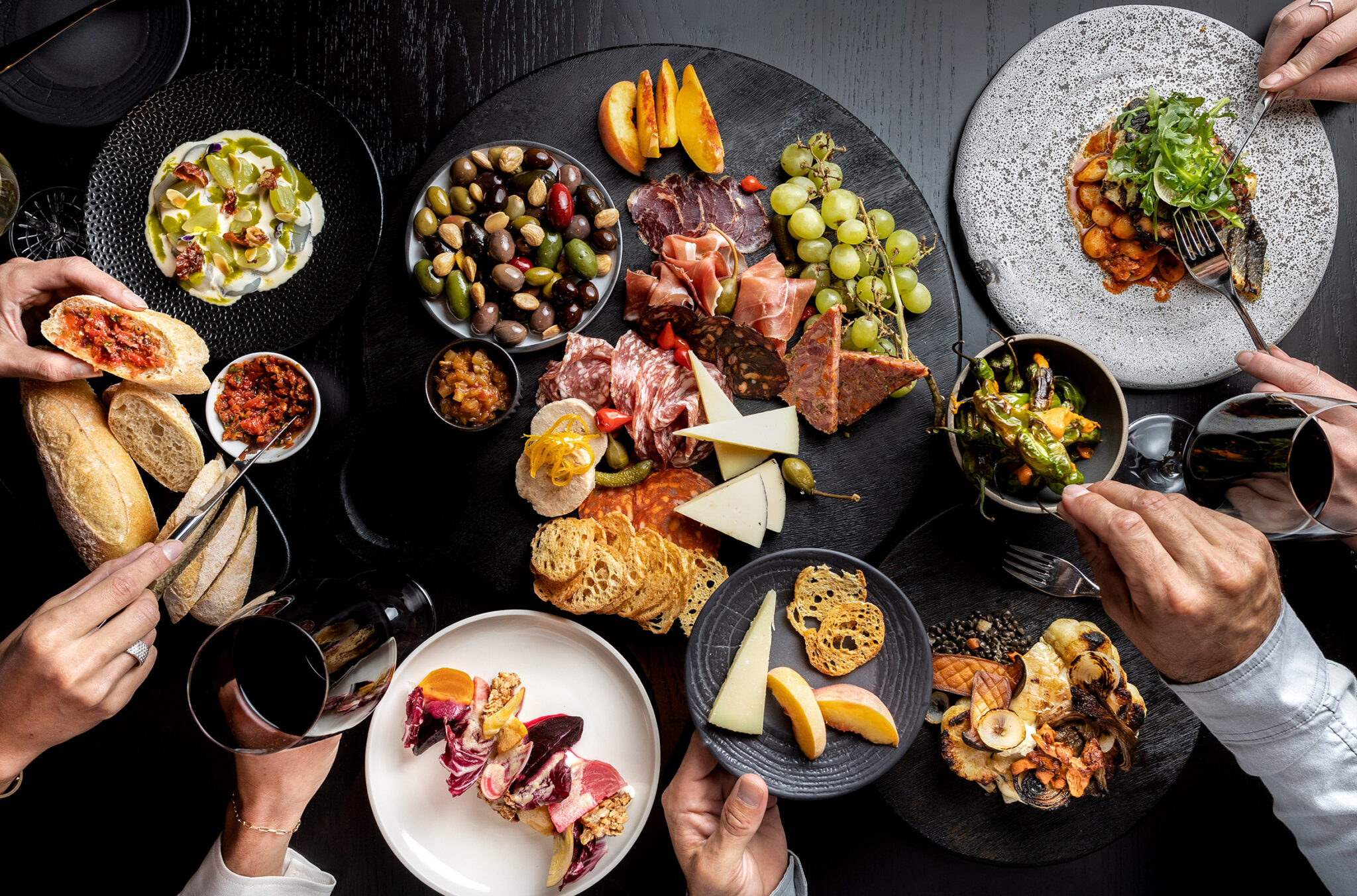 A beautiful and colourful array of food laid out on a dark table with multiple hands reaching in to take a bite.