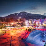 A shot of Whistler Olympic Plaza at nighttime in the winter. There are beautiful lights around the outdoor ice rink and trees, with Whistler Mountain in the background.