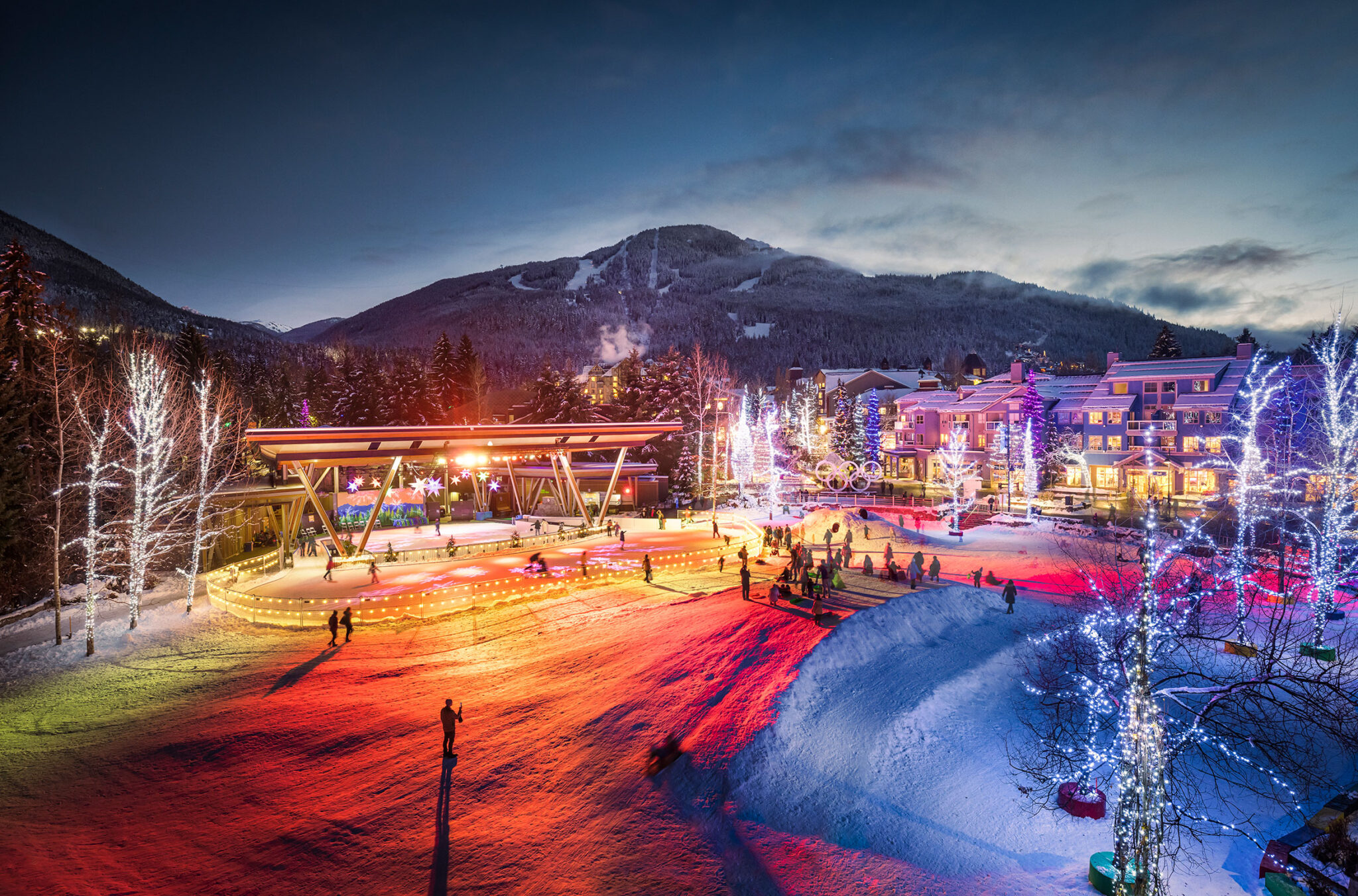 A shot of Whistler Olympic Plaza at nighttime in the winter. There are beautiful lights around the outdoor ice rink and trees, with Whistler Mountain in the background.