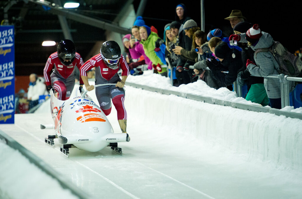 Two Canadian athletes run down the track at the Whistler Sliding Centre just before they jump in. A crowd cheers them on.