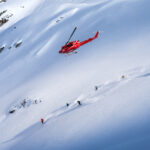 Heli skiers head down powdery slopes with a helicopter flying above them.