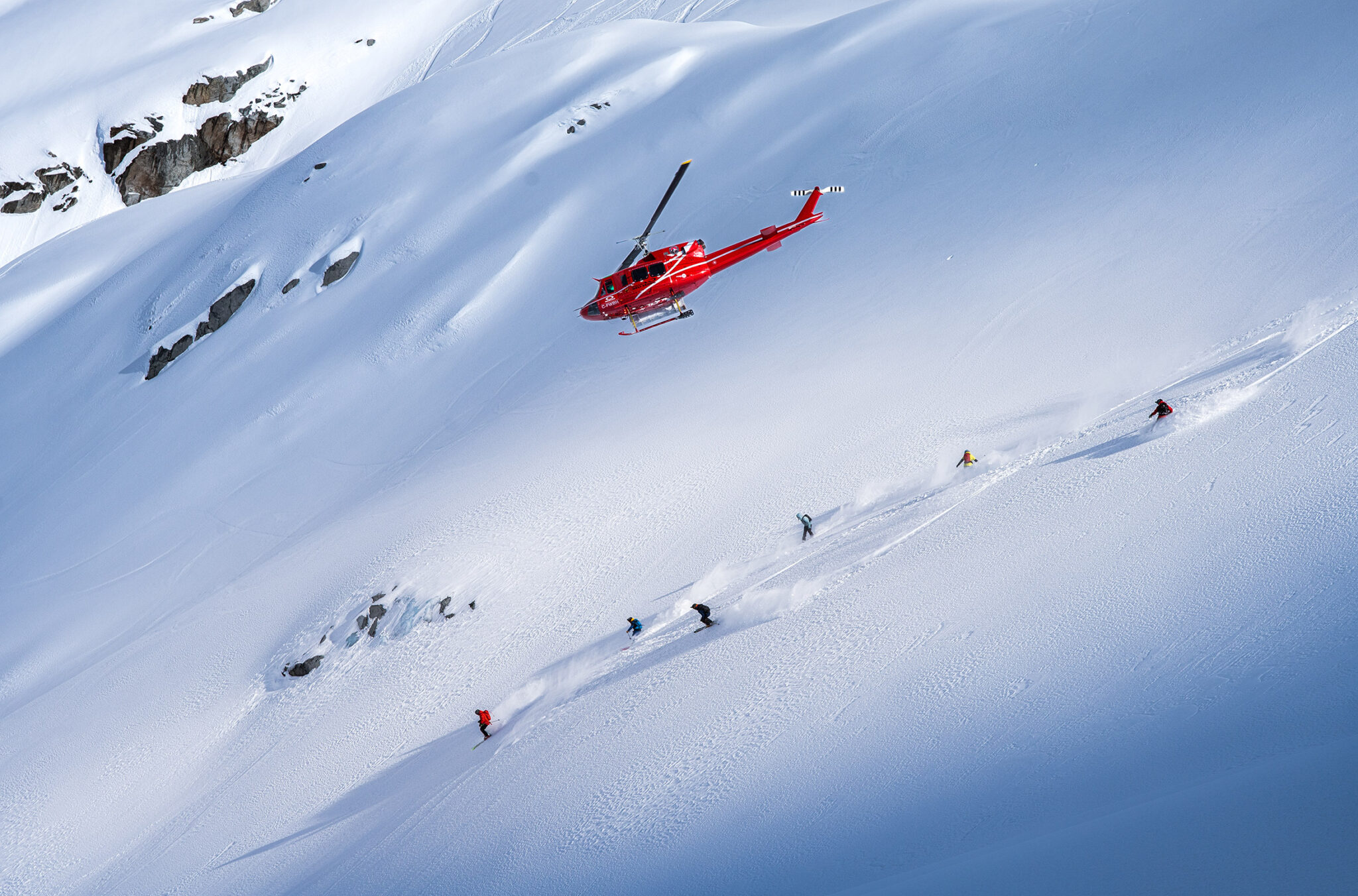 Heli skiers head down powdery slopes with a helicopter flying above them.