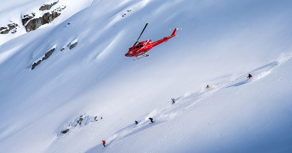 Heli skiers head down powdery slopes with a helicopter flying above them.