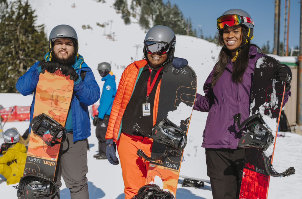 Three snowboarders face the camera smiling on an instruction day with Colour the Trails.
