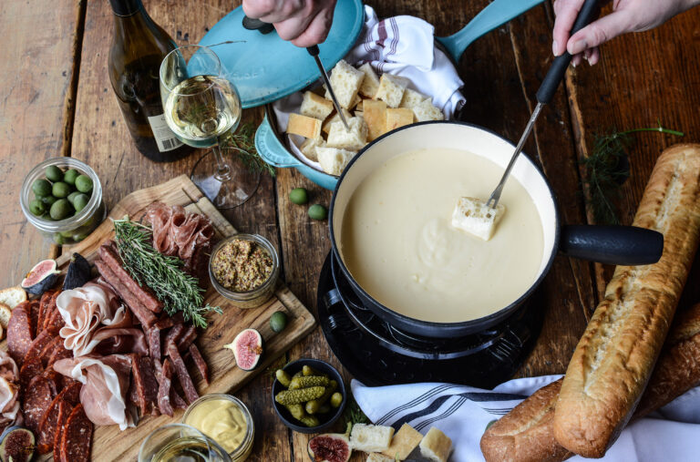 Someone reaching into a cheese fondue pot on a table laden with delicious items.