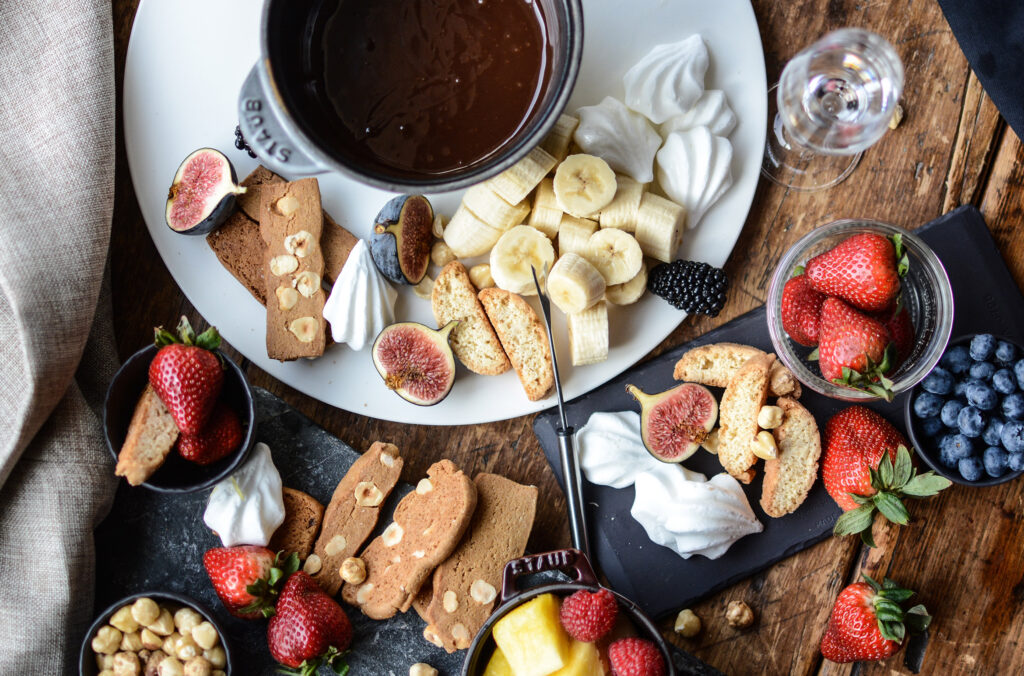 Someone reaching into a chocolate fondue pot on a table laden with delicious items.