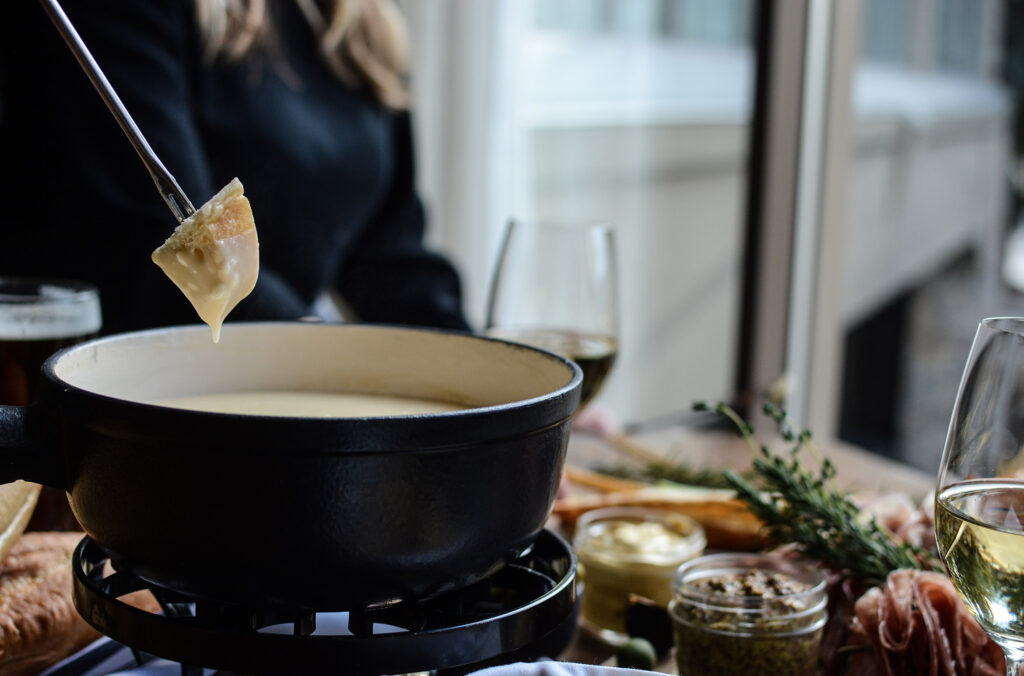 Someone reaching into a cheese fondue pot on a table laden with delicious items.