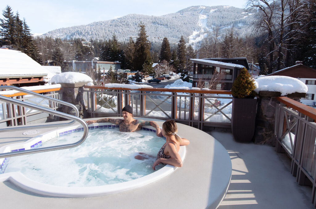 Two people enjoy Nita Lake Lodge's rooftop hot tub in the winter overlooking the mountains.