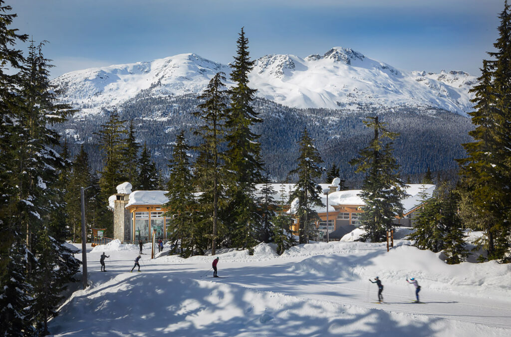 A shot of the Day Lodge at Whistler Olympic Park with the Coast Mountains in the background and cross-country skiers in the foreground.