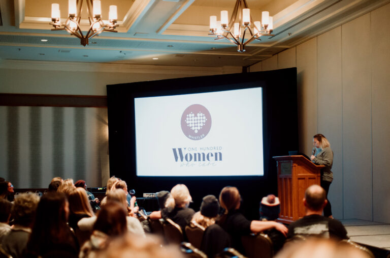 Ashlie stands at the front of the stage to start a 100 Women Who Care Whistler meeting.