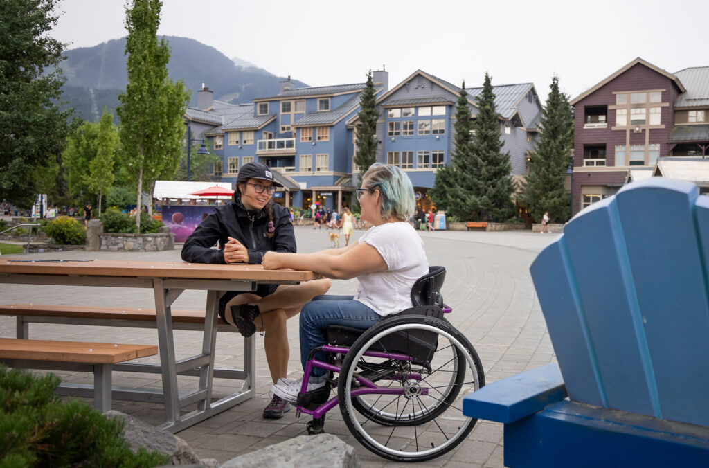 Two people seated at a wooden outdoor table in Whistler Olympic Plaza, engaging in conversation. The plaza is surrounded by modern mountain architecture and various pedestrians in a casual, relaxed atmosphere.