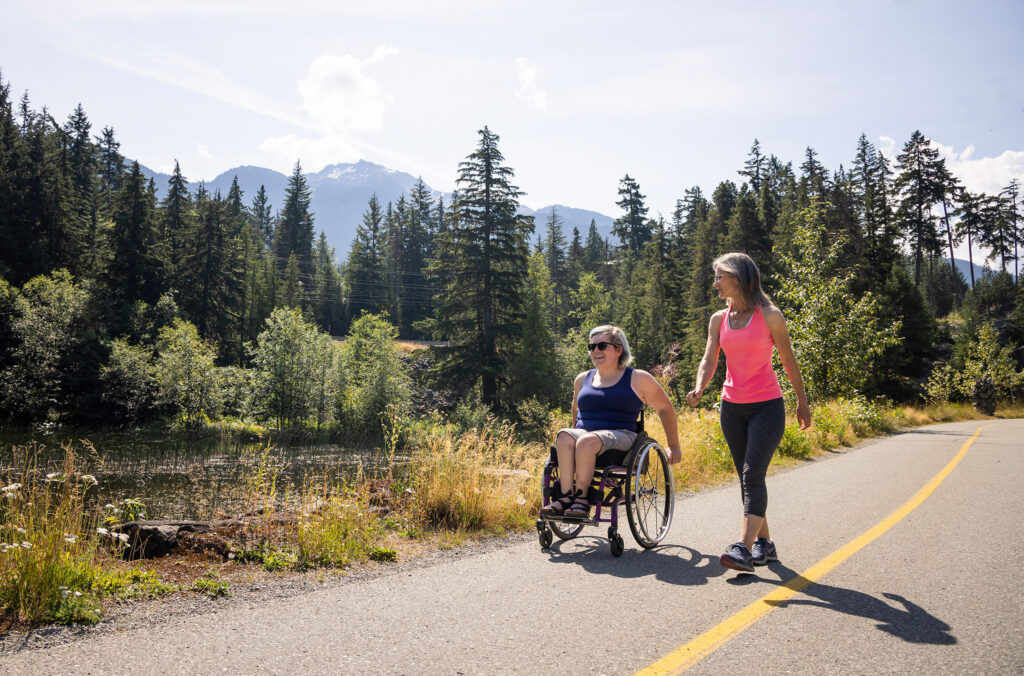 A sunny, tree-lined path in Whistler with two people enjoying a leisurely stroll in the summer sun.
