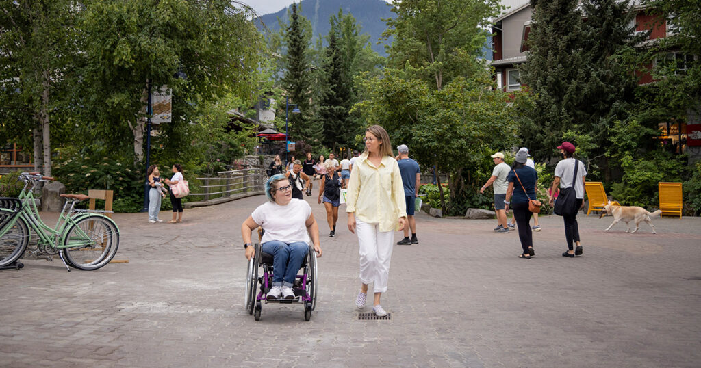 A lively pedestrian street in Whistler Village with a person in a wheelchair accompanied by another person walking beside her. The street is bustling with tourists and locals, surrounded by quaint shops and greenery.