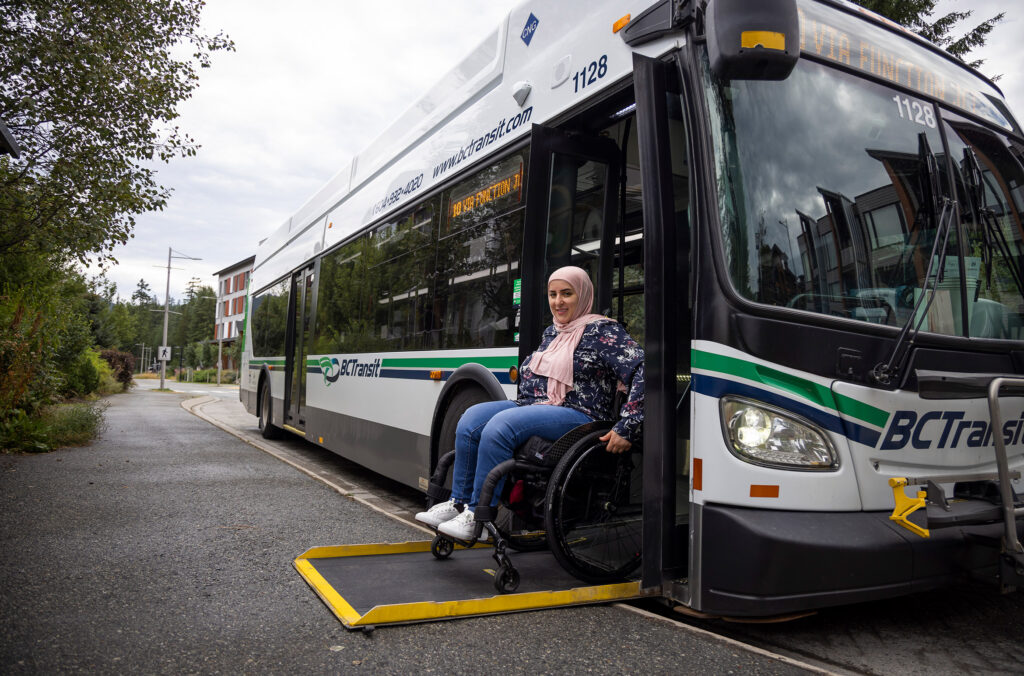 A person in a wheelchair smiling as they board a public bus via a yellow ramp in Whistler. The bus is parked in a lush, green setting with modern buildings in the background, under a cloudy sky.