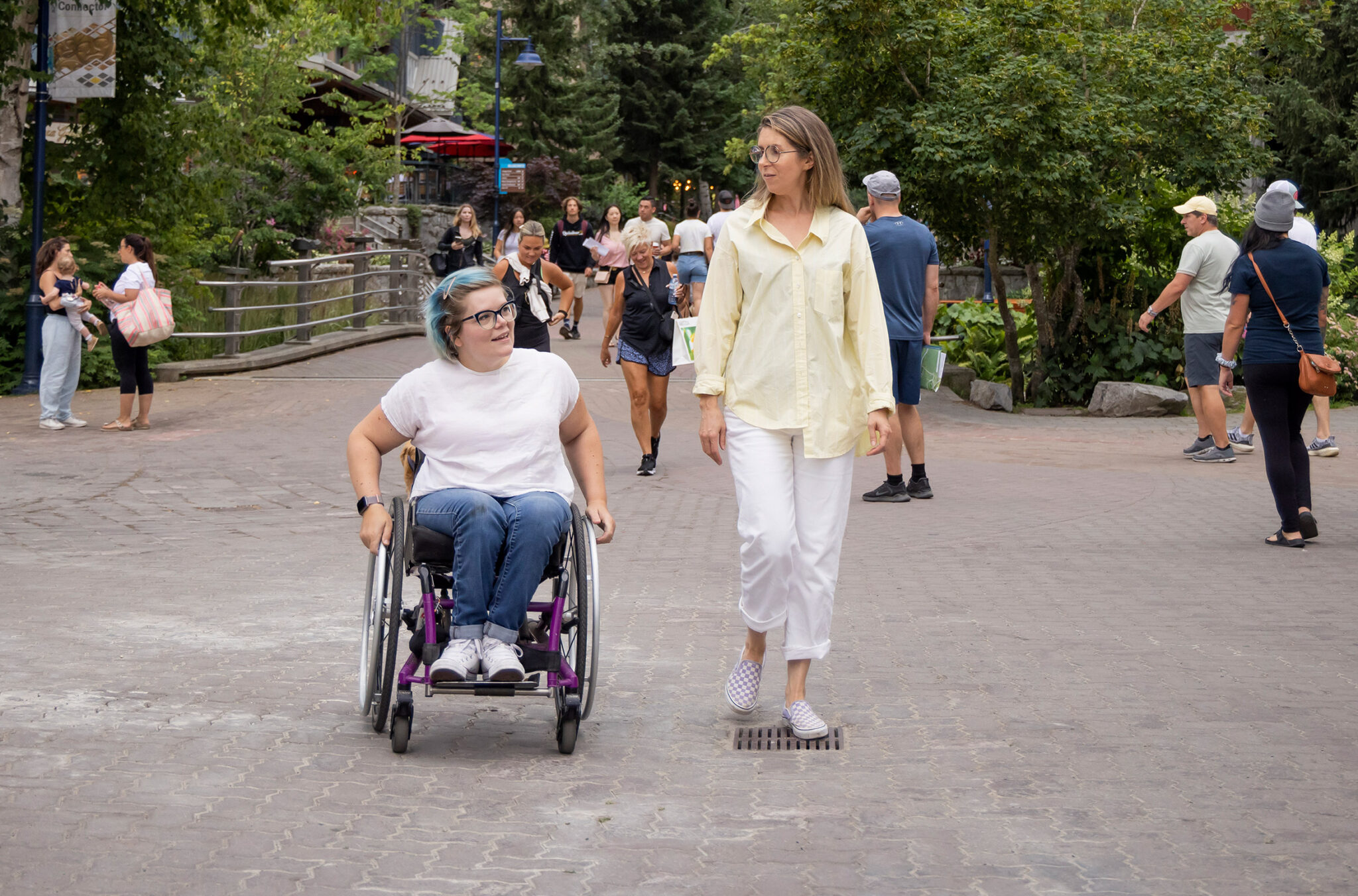 Two people enjoy the lively pedestrian Village Stroll in Whistler Village. The street is bustling with tourists and locals, surrounded by quaint shops and greenery.