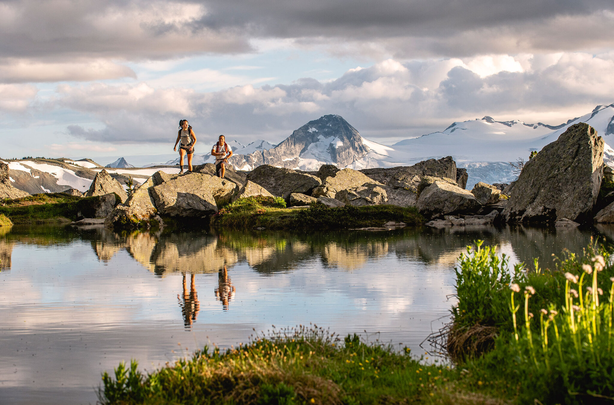 Two people hike in the high alpine on Whistler Blackcomb in the summer. There's a lake in front of them reflecting them as they hike.