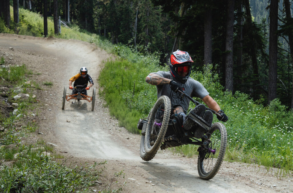 Two adaptive mountain bike riders enjoy pulling tricks as they make their way down the trails of the Whistler Mountain Bike Park.